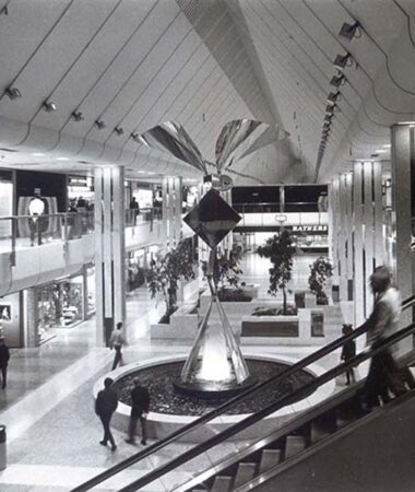 A grey scale photograph of a shopping centre in Harlow. A person is going down the escalator and other people walk around the fountain central to the shopping centre.