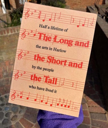 A photograph of someone holding a book named 'The Long and The Short and The Tall: Half a lifetime of the arts in Harlow by the people who have lived it' the book has music notes on it and is orange.