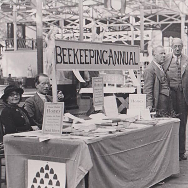A sepia-toned old photograph of a Beekeeping Annual stand, a few people are sat down at the stand and a couple gather around it.