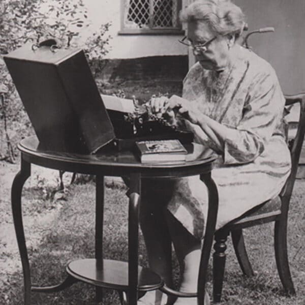 A black and white photograph of a woman sat at a table in a garden typing away on an old typewriter.